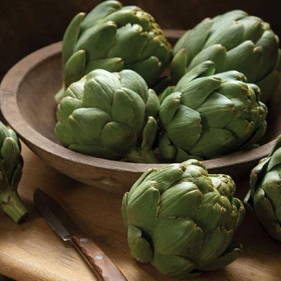 A wooden bowl filled with fresh artichokes on a wooden surface, with a knife partially visible to the side.