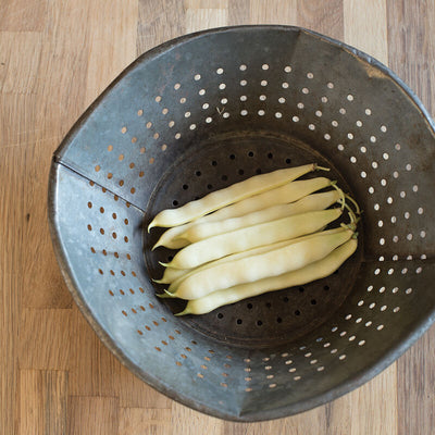A group of yellow Bean Bush Capitano beans laid out in a colander, which is placed on a wooden surface.