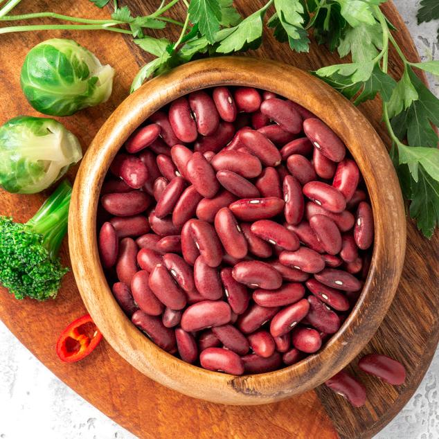 A wooden bowl filled with dry red kidney beans on a wooden surface, with parsley leaves, green beans, and a red pepper in the background.
