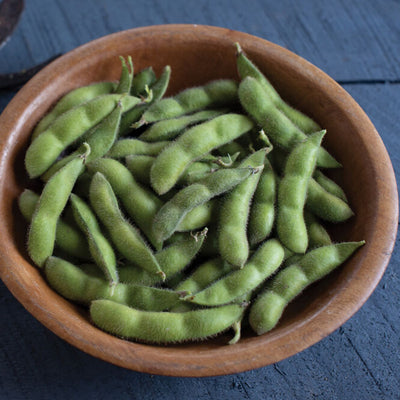 A wooden bowl filled with fresh green soybeans, with a pair of red gardening shears beside it on a dark wooden surface.