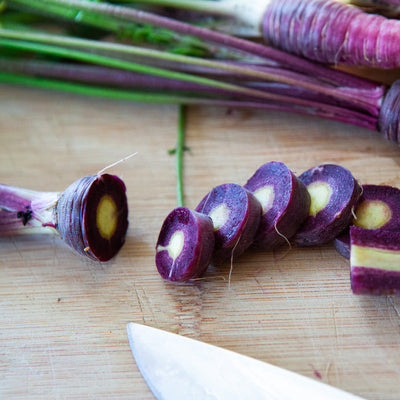 A cluster of dark purple carrot roots with one of them cut in half to show the interior, placed on a wooden surface with a knife beside them.