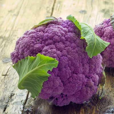 A fresh head of Cauliflower Purple of Sicily with vibrant purple florets and green leaves on a wooden surface.