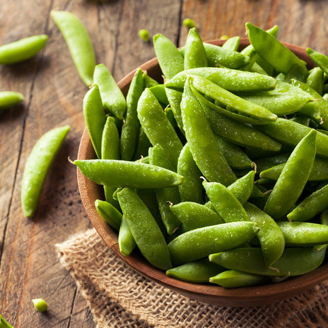 A wooden bowl filled with fresh green Sugar Snap peas on a rustic wooden table.