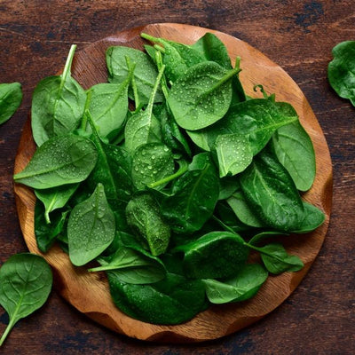 A bunch of fresh green spinach leaves on a wooden plate, with more leaves visible in the background.