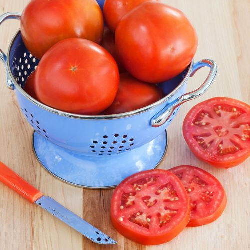 Tomatoes in a blue colander with sliced tomatoes on a wooden surface