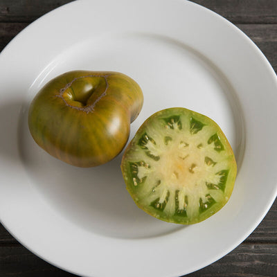 A whole Cherokee Green Tomato and a half slice of the same tomato on a white plate, displaying its green interior with a hint of orange.