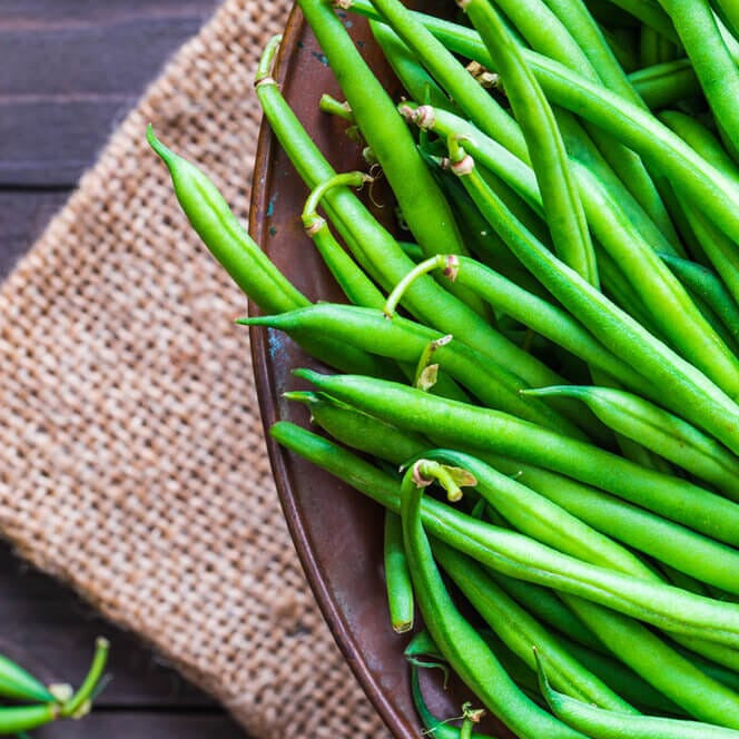A bunch of fresh green beans on a brown background, with a few beans placed on a burlap mat beside the bowl.