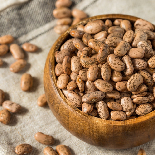 A wooden bowl filled with dried pinto beans, with more beans scattered around on a linen surface.
