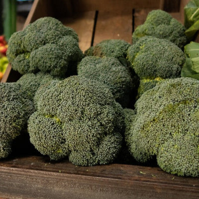 A wooden crate filled with fresh broccoli on a market table, with a blurred background of various fruits.