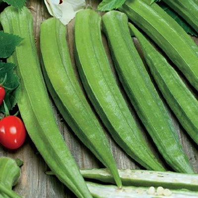 A cluster of long, slender green okra pods on a wooden surface, with a few tomatoes visible to the side.