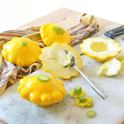 A group of yellow summer squashes, both whole and sliced, with a spoonful of cream cheese on one of the squashes, placed on a marble surface with a striped cloth in the background.