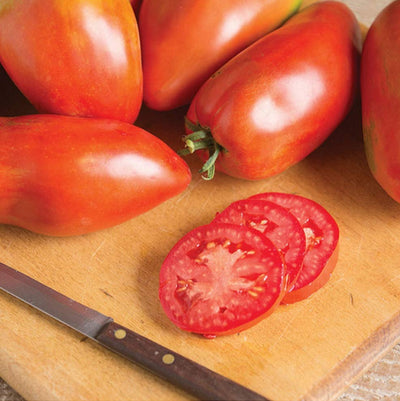 A knife with a slice of tomato on it, placed on a wooden cutting board with whole tomatoes in the background.
